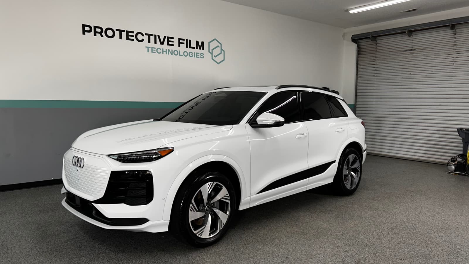 A white Audi SUV is parked indoors on a gray carpet near a wall with "Protective Film Technologies" signage and a partially open metal garage door.