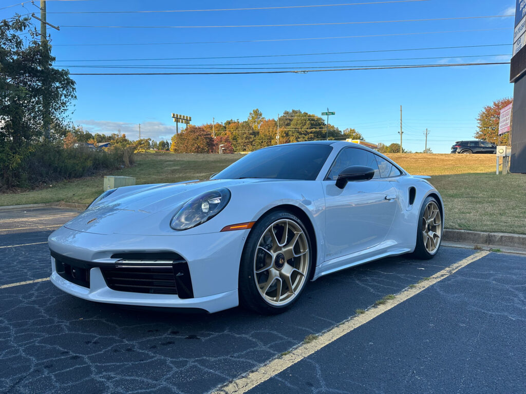 A white sports car with tinted windows and gold wheels is parked in a parking lot on a sunny day.
