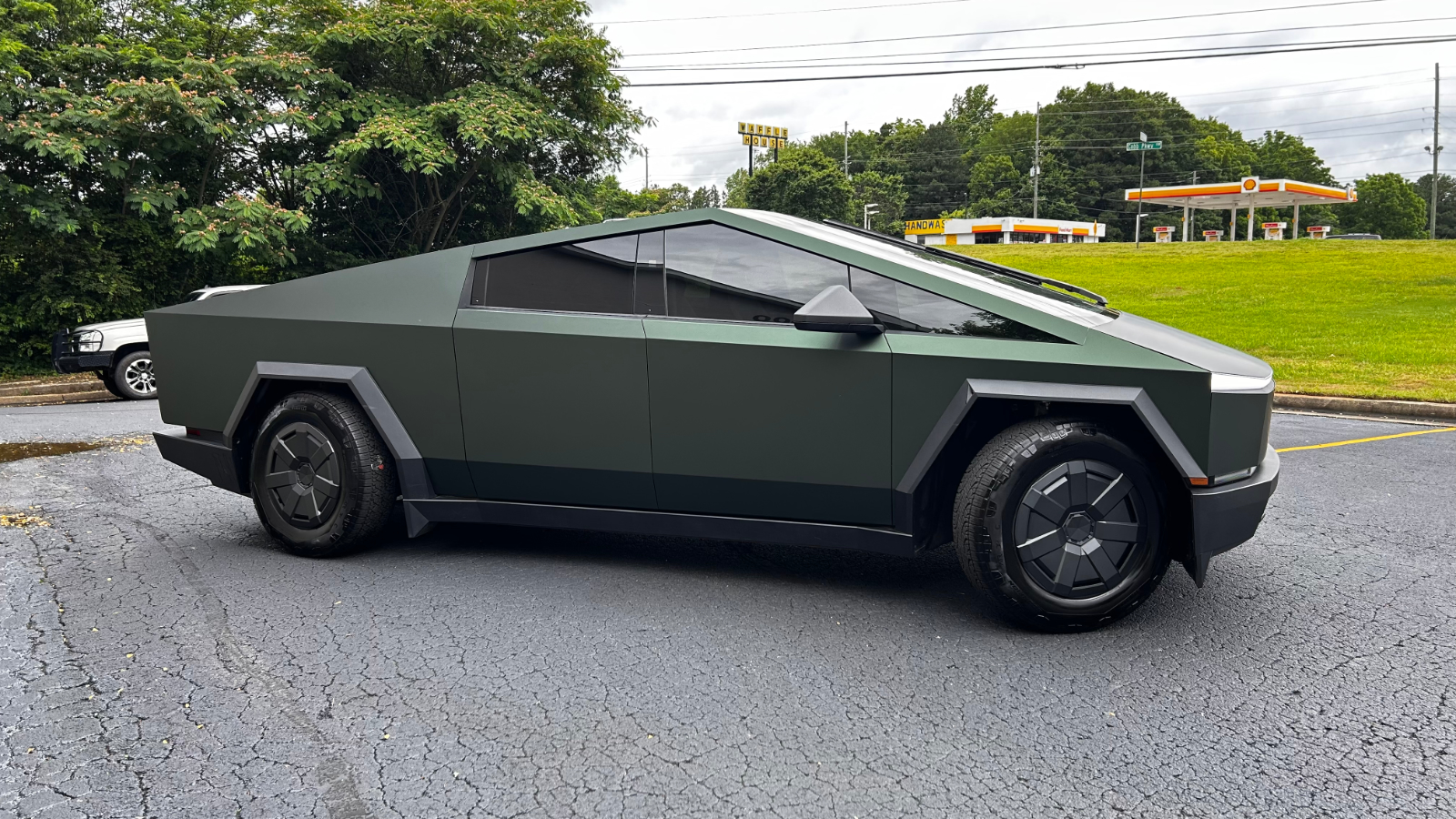 A matte green Tesla Cybertruck is parked on asphalt near a grassy area with trees and a gas station in the background.