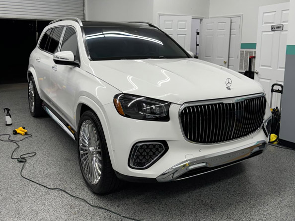 A white Mercedes-Benz SUV is parked indoors in a garage or detailing shop, with cleaning equipment visible nearby.