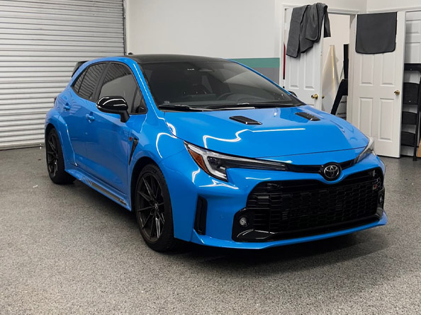 A blue Toyota GR Corolla hatchback is parked indoors on a gray speckled floor near a white wall and open doors with towels hanging.