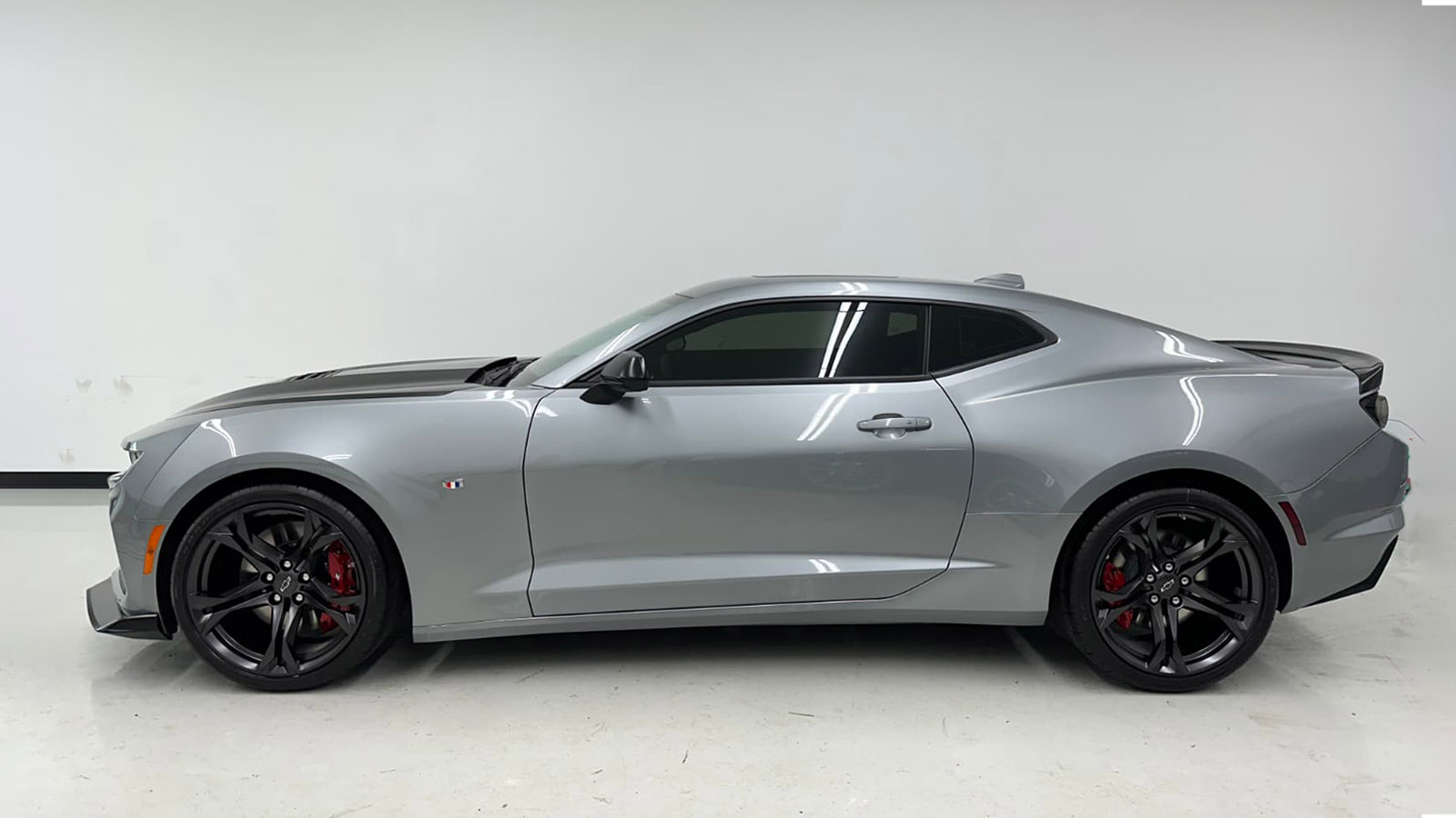 A silver two-door sports car with tinted windows and black wheels is parked indoors against a plain white wall.